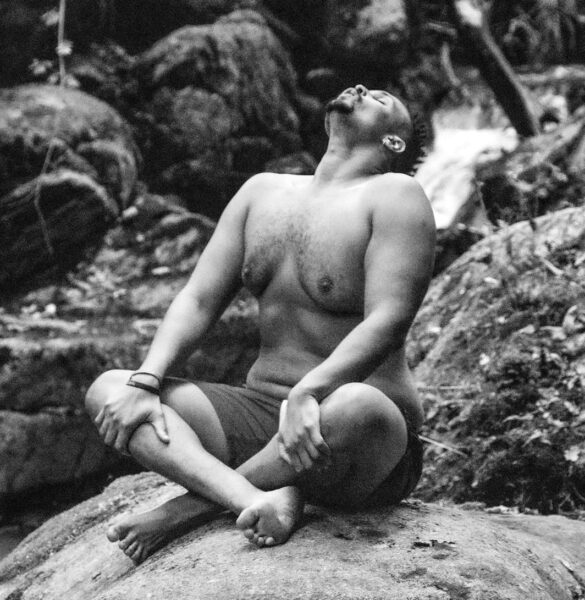 Man sitting cross-legged on a rock by a stream with head tilted up (black-and-white photo)